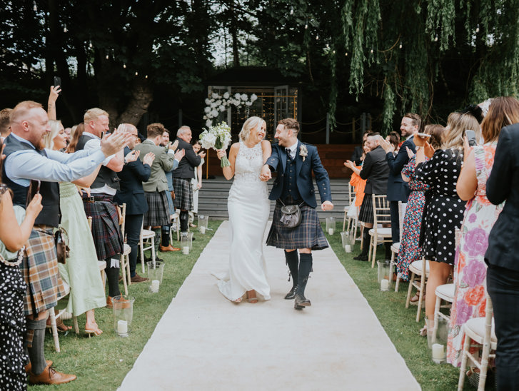 Bride and groom smile together as they walk back down the aisle at The Venue, Eskmills while their guests clap